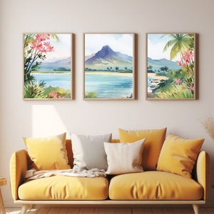May include: A set of three framed watercolour prints depicting a tropical coastal landscape with a central mountain, blue water, sandy beaches, and pink flowers. The artwork is displayed in light wood frames on a plain wall above a yellow sofa.