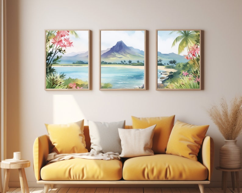 May include: A set of three framed watercolour prints depicting a tropical coastal landscape with a central mountain, blue water, sandy beaches, and pink flowers. The artwork is displayed in light wood frames on a plain wall above a yellow sofa.