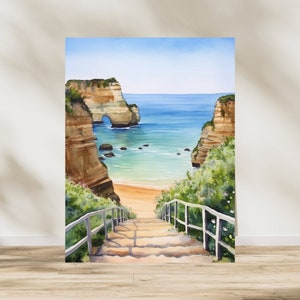 May include: Watercolor painting of a stone staircase leading down to a beach with a rock formation in the background. The beach is surrounded by cliffs and the ocean is a light blue color.