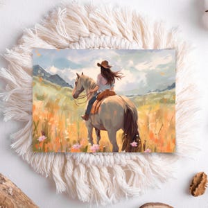 May include: A watercolour print shows a person riding a horse through a field of wildflowers. The rider wears a brown hat and a light-coloured shirt. The horse is light grey, and the background features mountains and a cloudy sky. The print is set on a white, fringed placemat.