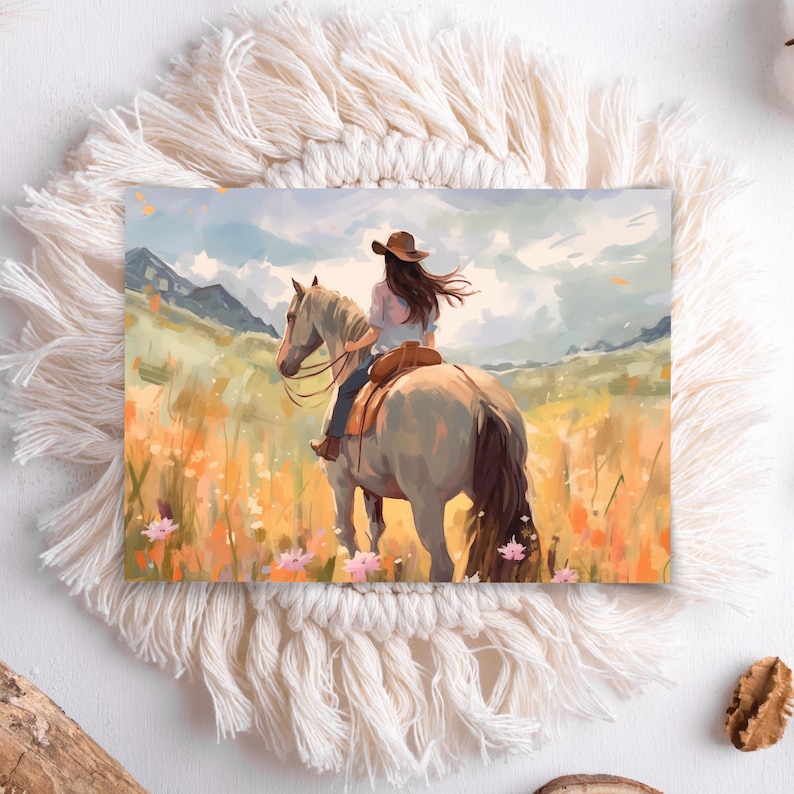 May include: A watercolour print shows a person riding a horse through a field of wildflowers. The rider wears a brown hat and a light-coloured shirt. The horse is light grey, and the background features mountains and a cloudy sky. The print is set on a white, fringed placemat.