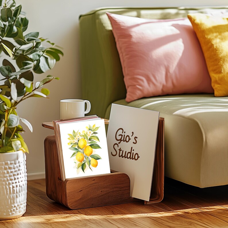May include: A dark wood magazine rack holding a white card with a watercolor illustration of yellow lemons and green leaves, alongside a white card featuring the brown text Gio's Studio, positioned next to a white ceramic mug.