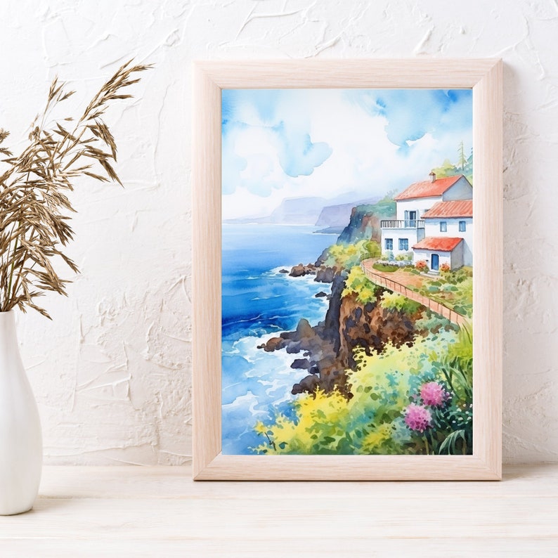 May include: Watercolor painting of a coastal scene with white buildings, red roofs, and a pathway along a cliff. The artwork is framed in a light wood frame, with a blue ocean and sky in the background. The painting is displayed on a white surface.