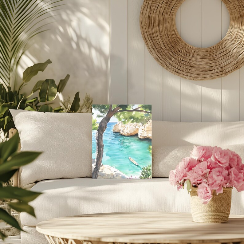 May include: Watercolor painting of a coastal scene with turquoise water, cliffs, and a small boat. A tree frames the view, with green foliage and a clear blue sky. The painting is displayed on a white sofa with a woven basket of pink flowers.