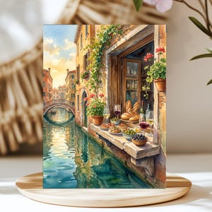 May include: A watercolor painting of a Venetian canal scene featuring a stone ledge with wine glasses, bowls of olives, and loaves of bread. A bridge spans the water in the background, while climbing vines and red flowers decorate the building facade.