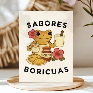 May include: A greeting card featuring a cartoon frog wearing an apron and holding a coffee cup, a glass of coquito, and a jar of sofrito. The card has the words "Sabores Boricuas" in black letters, with a hibiscus flower on the frog's head and another one on the bottom right.