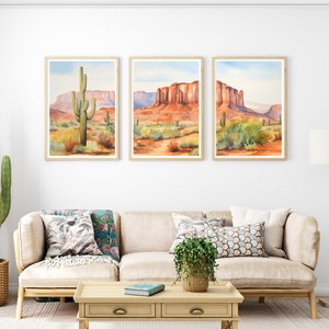 May include: A triptych of framed watercolour landscape prints featuring desert scenes with red rock formations, mesas, and saguaro cacti under a pale blue sky. The artwork is displayed in light wood frames on a white wall above a beige sofa.