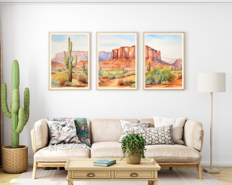 May include: A triptych of framed watercolour landscape prints featuring desert scenes with red rock formations, mesas, and saguaro cacti under a pale blue sky. The artwork is displayed in light wood frames on a white wall above a beige sofa.