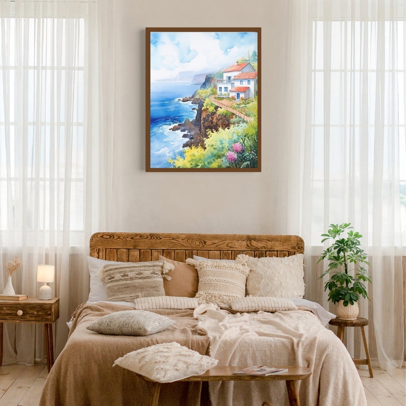 May include: A framed watercolor painting depicts a coastal scene with a white building, red roof, and a pathway along a cliff. The artwork features vibrant blues, greens, and yellows, with a wooden frame. The painting is displayed above a bed with neutral-toned bedding.