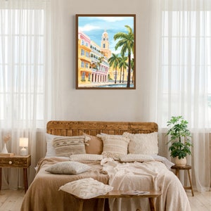 Peut inclure: Une peinture &agrave; l'aquarelle encadr&eacute;e repr&eacute;sentant une sc&egrave;ne de rue de ville avec des b&acirc;timents jaunes, des palmiers et un ciel bleu. La peinture est accroch&eacute;e au mur au-dessus d'un lit en bois avec une couverture beige et des oreillers.