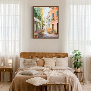 May include: A watercolor painting of a narrow street in a European city. The buildings are painted in shades of yellow, pink, and blue, and there are flowers in pots along the street. The painting is framed in a brown wooden frame.