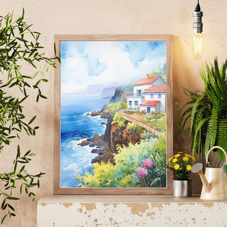 May include: Watercolor painting of a coastal scene with white houses, red roofs, and a pathway along a cliff. The artwork is framed in a light wood frame. The ocean is a deep blue, and the sky is a light blue with fluffy clouds. Flowers and greenery are in the foreground.