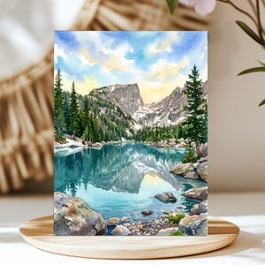 May include: A watercolor painting depicting a mountain lake scene with a jagged peak reflecting in the turquoise water. The landscape features dense green pine trees, rocky shorelines, and a soft sky with yellow and blue hues above the mountains.