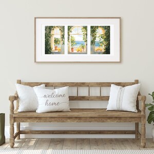 May include: A wooden bench with two pillows, one with the text "welcome home" and the other with white and gray stripes. A green plant in a white pot is on the right side of the bench. A framed print of three windows with a view of the ocean and lemon trees is hanging on the wall behind the bench.