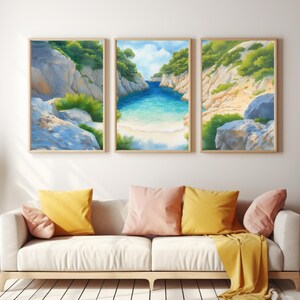 May include: Three framed landscape art prints depicting a coastal scene with turquoise water, sandy beaches, and rocky cliffs. The artwork is in a light, pastel color palette. The prints are displayed above a cream-colored sofa with yellow and pink pillows.