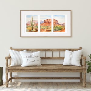May include: A triptych watercolour print in a light wood frame depicting a desert landscape with red rock formations and a saguaro cactus. Below, a wooden bench holds two white pillows, one with the text welcome home, and two green boots.