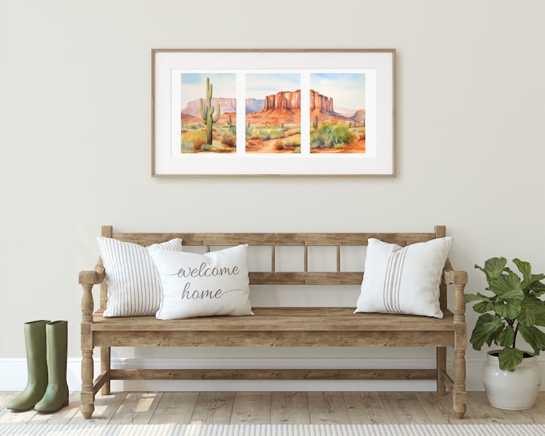 May include: A triptych watercolour print in a light wood frame depicting a desert landscape with red rock formations and a saguaro cactus. Below, a wooden bench holds two white pillows, one with the text welcome home, and two green boots.