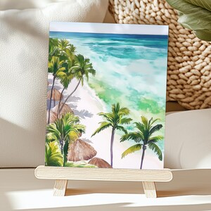 Puede incluir: Pintura a la acuarela de una escena de playa tropical. La obra de arte representa una playa de arena blanca, agua turquesa, palmeras y sombrillas de paja. La pintura se exhibe en un peque&ntilde;o caballete de madera.