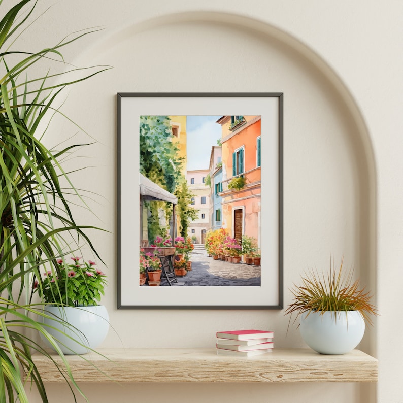 May include: A watercolor painting of a narrow street in a European city. The street is lined with colorful buildings and potted plants. The painting is framed in a gray frame and is hanging on a white wall.