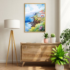 May include: Watercolor painting of a coastal scene with a white building, red roof, and blue doors. The artwork is framed in a gold-colored frame. The painting depicts a cliffside with lush greenery, pink flowers, and the ocean. The artwork is displayed above a wooden cabinet.