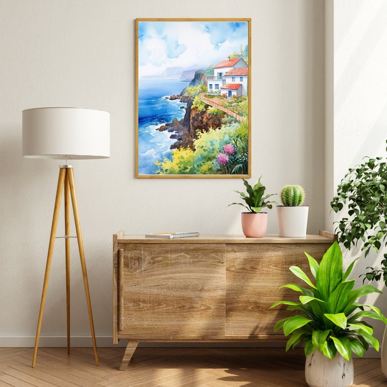 May include: Watercolor painting of a coastal scene with a white building, red roof, and blue doors. The artwork is framed in a gold-colored frame. The painting depicts a cliffside with lush greenery, pink flowers, and the ocean. The artwork is displayed above a wooden cabinet.