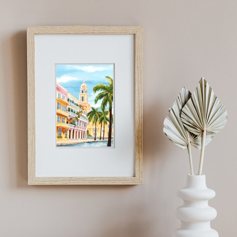 Peut inclure: Une peinture &agrave; l'aquarelle encadr&eacute;e d'un b&acirc;timent color&eacute; avec une haute tour et des palmiers devant. Le b&acirc;timent est jaune avec des accents roses et bleus. Les palmiers sont verts et le ciel est bleu.