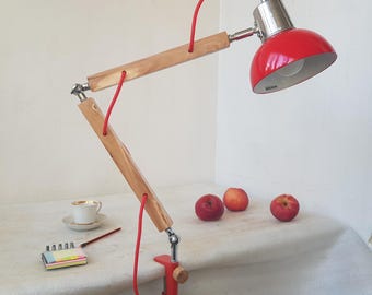 May include: A red desk lamp with a wooden arm and a red cord. The lamp has a silver head and is clamped to a surface. Three red apples, a teacup, a pencil, and a notepad are also on the surface.