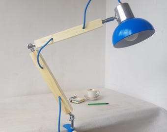 May include: A blue and silver desk lamp with a wooden arm and clamp base. The lamp has a blue lampshade and a flexible arm. The lamp is attached to a table with a clamp. A cup of coffee, a notepad, and a pencil are on the table.