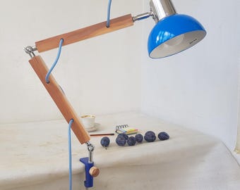May include: A blue and silver desk lamp with a wooden arm and a blue cord. The lamp is clamped to a white surface. A cup of tea, a pencil, and plums are on the surface.