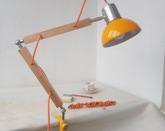 May include: A yellow and silver desk lamp with a wooden arm and orange cord. The lamp has a clamp base and a yellow lampshade. The lamp is adjustable and has a modern design.