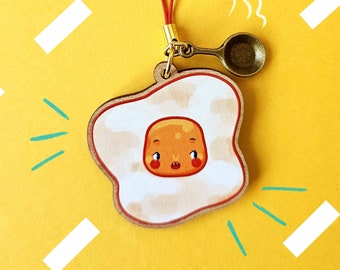 May include: A wooden charm featuring a cartoon fried egg with a smiling face. The charm is attached to a red cord and a small bronze spoon. The background is yellow with white diagonal lines and teal lines.