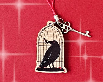 May include: A wooden charm featuring a black crow inside a cage, with a silver key attached. The charm hangs from a red cord. The crow holds a sprig in its beak. The background is red with white starbursts.