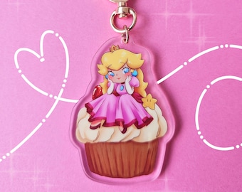 May include: A clear acrylic keychain featuring a cartoon illustration of Princess Peach from the Super Mario franchise. Peach is wearing a pink dress and a crown, and she is sitting on top of a cupcake. The keychain has a gold-colored metal clasp.