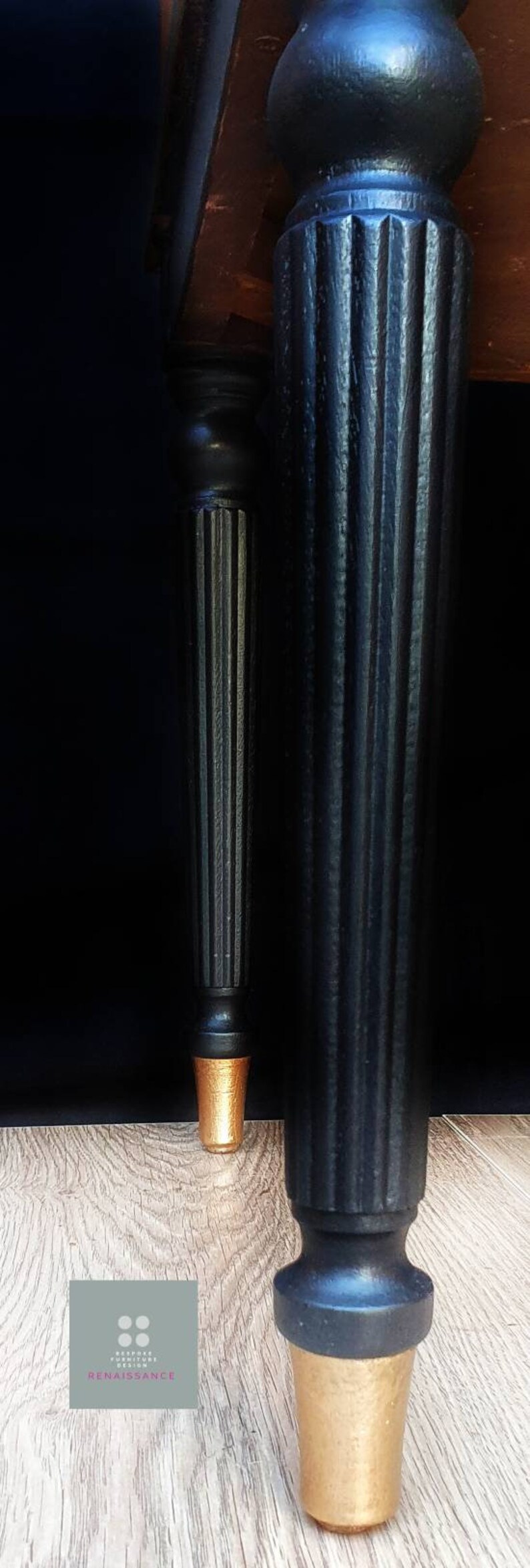 May include: Two black wooden table legs with gold-painted feet. The legs have a fluted design and are resting on a wooden floor.