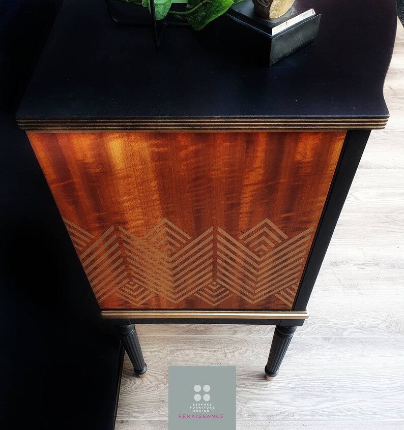 May include: A black and gold vintage cabinet with a wood grain finish and a geometric pattern on the front. The cabinet has black legs and a gold trim.