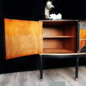 May include: A black and gold vintage cabinet with a geometric design on the door. The cabinet has two shelves and is sitting on a wooden floor.