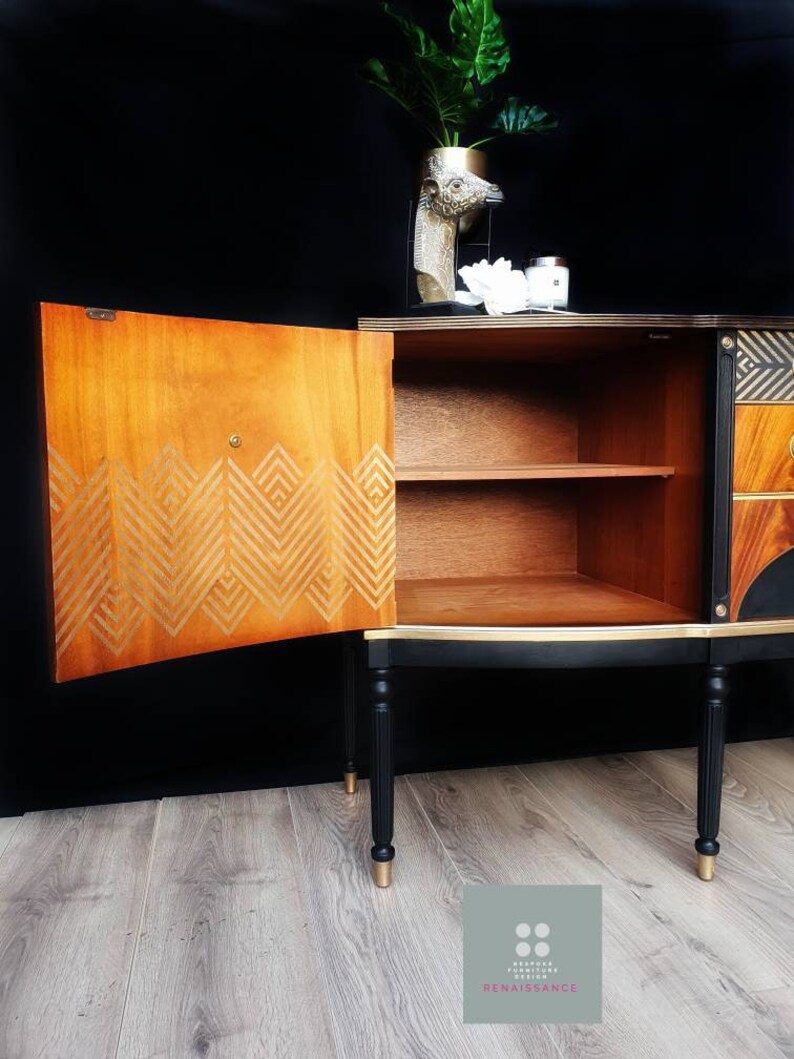 May include: A black and gold vintage cabinet with a geometric design on the door. The cabinet has two shelves and is sitting on a wooden floor.