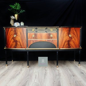 May include: A vintage wooden sideboard with black and gold accents. The sideboard has two doors on either side and a drawer in the center. The doors and drawer have gold handles. The sideboard is sitting on four black legs with gold tips. The sideboard is in front of a black background.