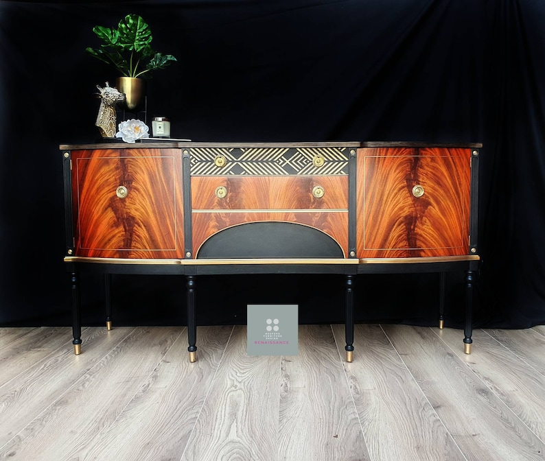 May include: A vintage wooden sideboard with black and gold accents. The sideboard has two doors on either side and a drawer in the center. The doors and drawer have gold handles. The sideboard is sitting on four black legs with gold tips. The sideboard is in front of a black background.