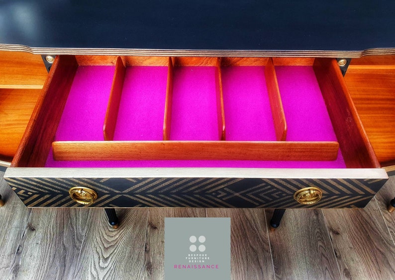 May include: A black and gold sideboard with a drawer that has a pink felt liner and wooden dividers. The drawer is open and the interior is visible. The sideboard is sitting on a wood floor.