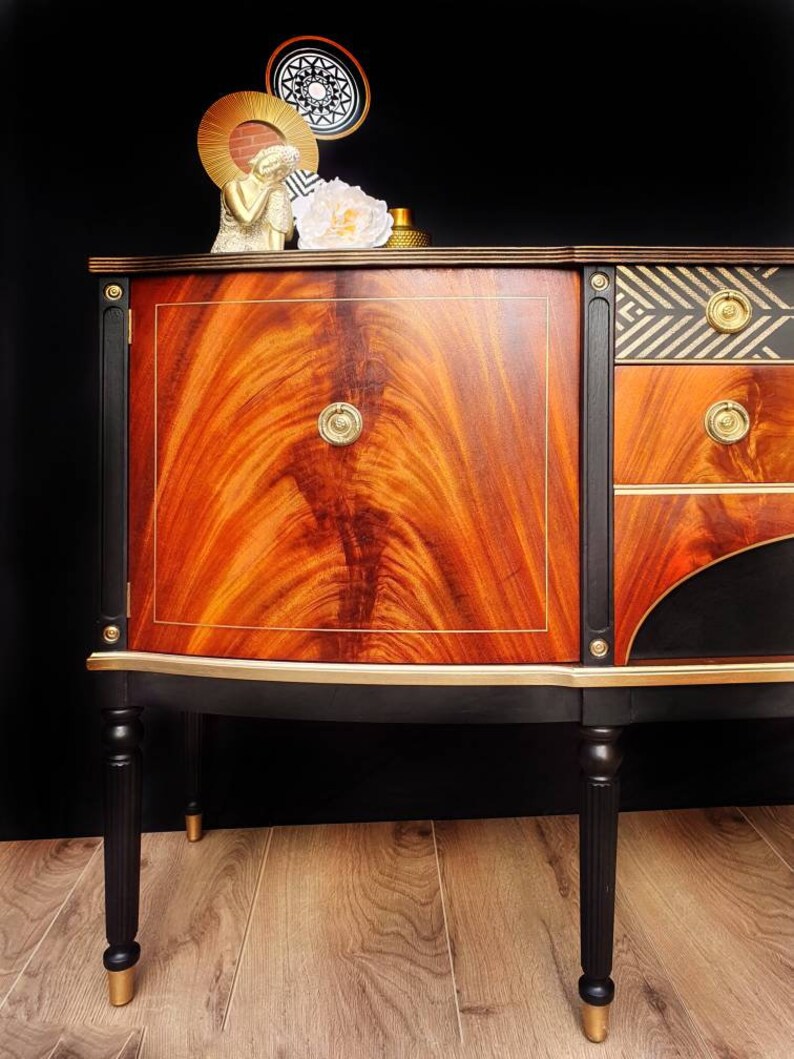 May include: A vintage wooden sideboard with black and gold painted details. The sideboard has a curved front and four legs. The top of the sideboard is decorated with a geometric pattern in black and gold.