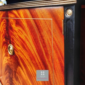 May include: A close-up of a wooden cabinet door with a rich, reddish-brown wood grain. The door has a gold-colored trim and a brass knob. The cabinet is painted black on the edges and has a gold-colored inscription that reads "BESPOKE FURNITURE DESIGN RENAISSANCE".