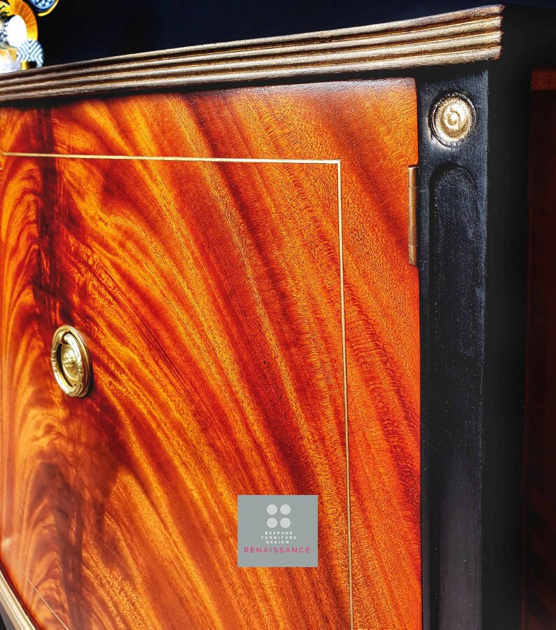 May include: A close-up of a wooden cabinet door with a rich, reddish-brown wood grain. The door has a gold-colored trim and a brass knob. The cabinet is painted black on the edges and has a gold-colored inscription that reads "BESPOKE FURNITURE DESIGN RENAISSANCE".