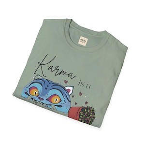 May include: Sage green t-shirt with the phrase "Karma is a" and a cartoon cat design. The cat is blue with yellow eyes and is peeking out from behind a red flower pot. The design includes small pink hearts and sparkles.