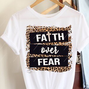 May include: White t-shirt with rolled-up sleeves. The shirt features a design with the words "FAITH over FEAR" in white, with a cross above "FAITH". The text is set against a black background with a leopard print border.