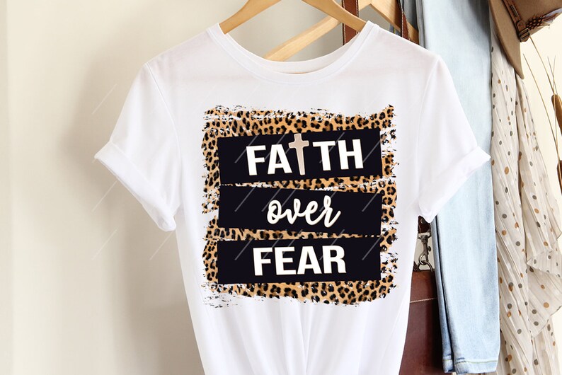 May include: White t-shirt with rolled-up sleeves. The shirt features a design with the words "FAITH over FEAR" in white, with a cross above "FAITH". The text is set against a black background with a leopard print border.