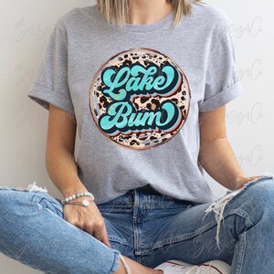 May include: A gray t-shirt featuring a circular design with the words "Lake Bum" in turquoise script. The design has a leopard print background and a copper-colored border. The shirt is worn by a person.