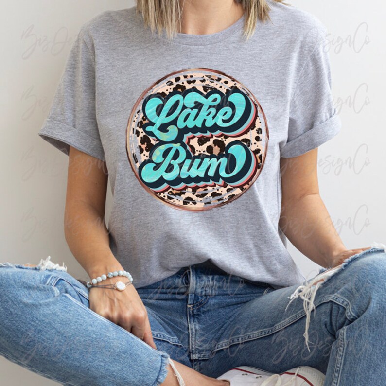 May include: A gray t-shirt featuring a circular design with the words "Lake Bum" in turquoise script. The design has a leopard print background and a copper-colored border. The shirt is worn by a person.