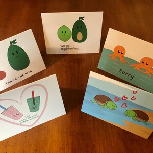 May include: Five greeting cards with different illustrations and text. The cards are on a wooden surface. The cards feature illustrations of avocados, turtles, and octopuses. The text on the cards includes "That's the pits", "We go together like...", "Sorry", and "You make me feel bubbly inside!"