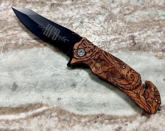 May include: A black folding knife with a wooden handle featuring intricate carved designs. The blade has the initials "HEB" etched into it. The knife is a tactical tool with a serrated edge and a pocket clip.